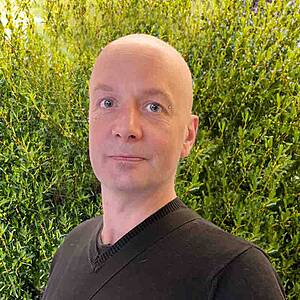 Bald man smiling against green foliage background.
